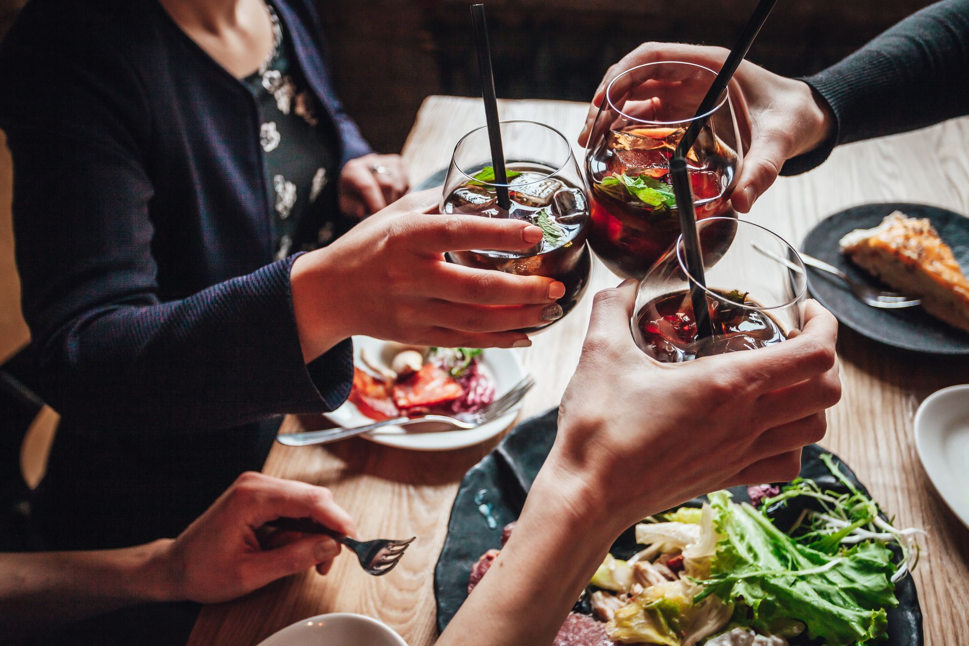 A group of people cheers with glasses of red drinks over a dinner table with salads and bread.