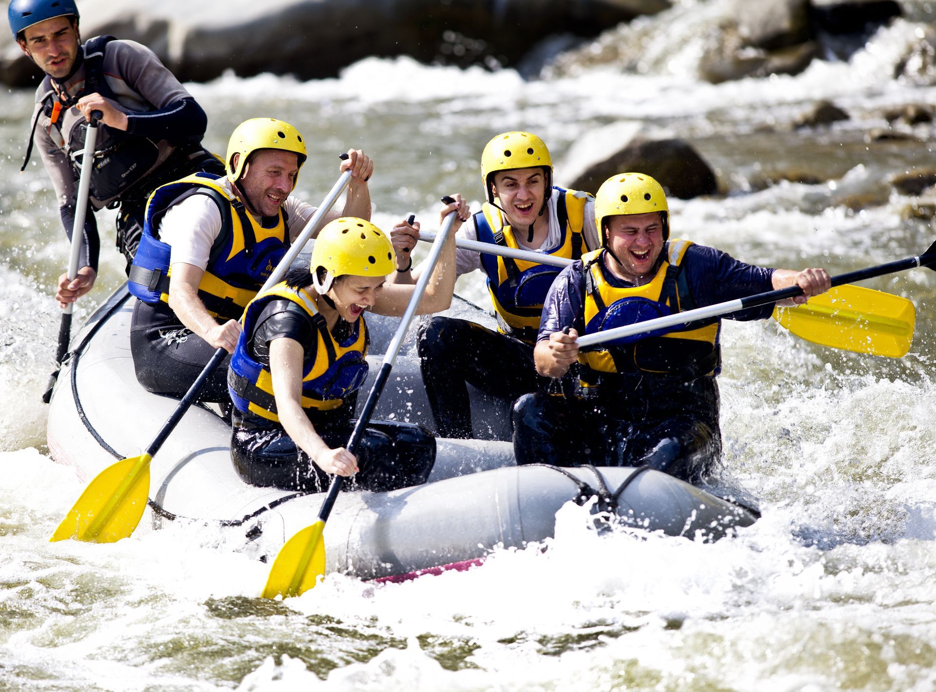 Four people in helmets and life vests whitewater rafting in a boat, actively paddling through river rapids.