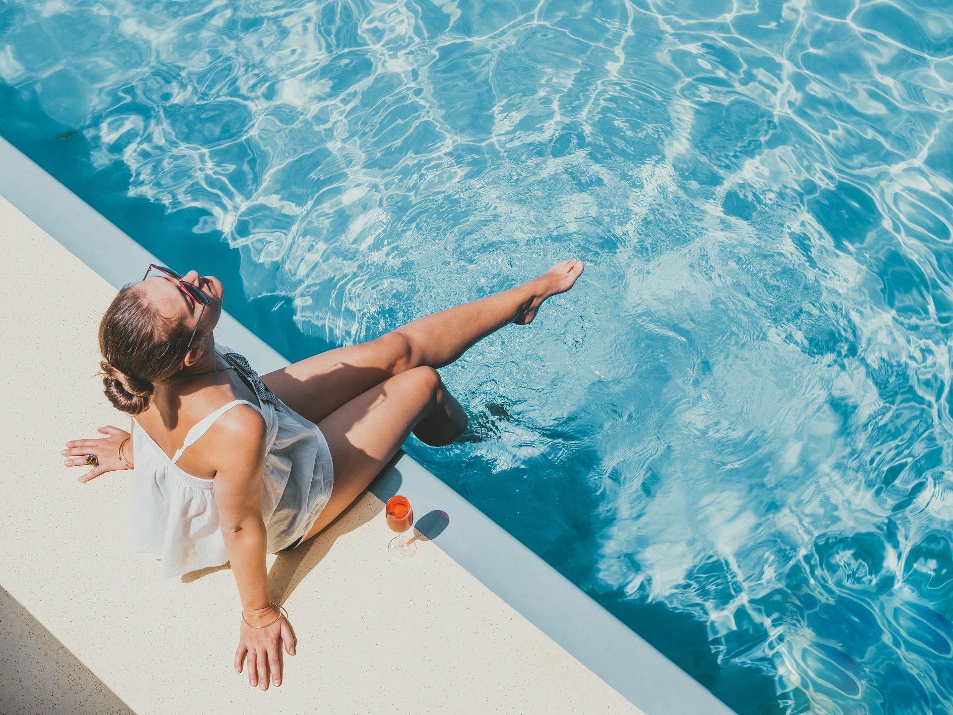 A person in a white top sits on a pool edge, dangling their feet in the bright blue water with a drink nearby.