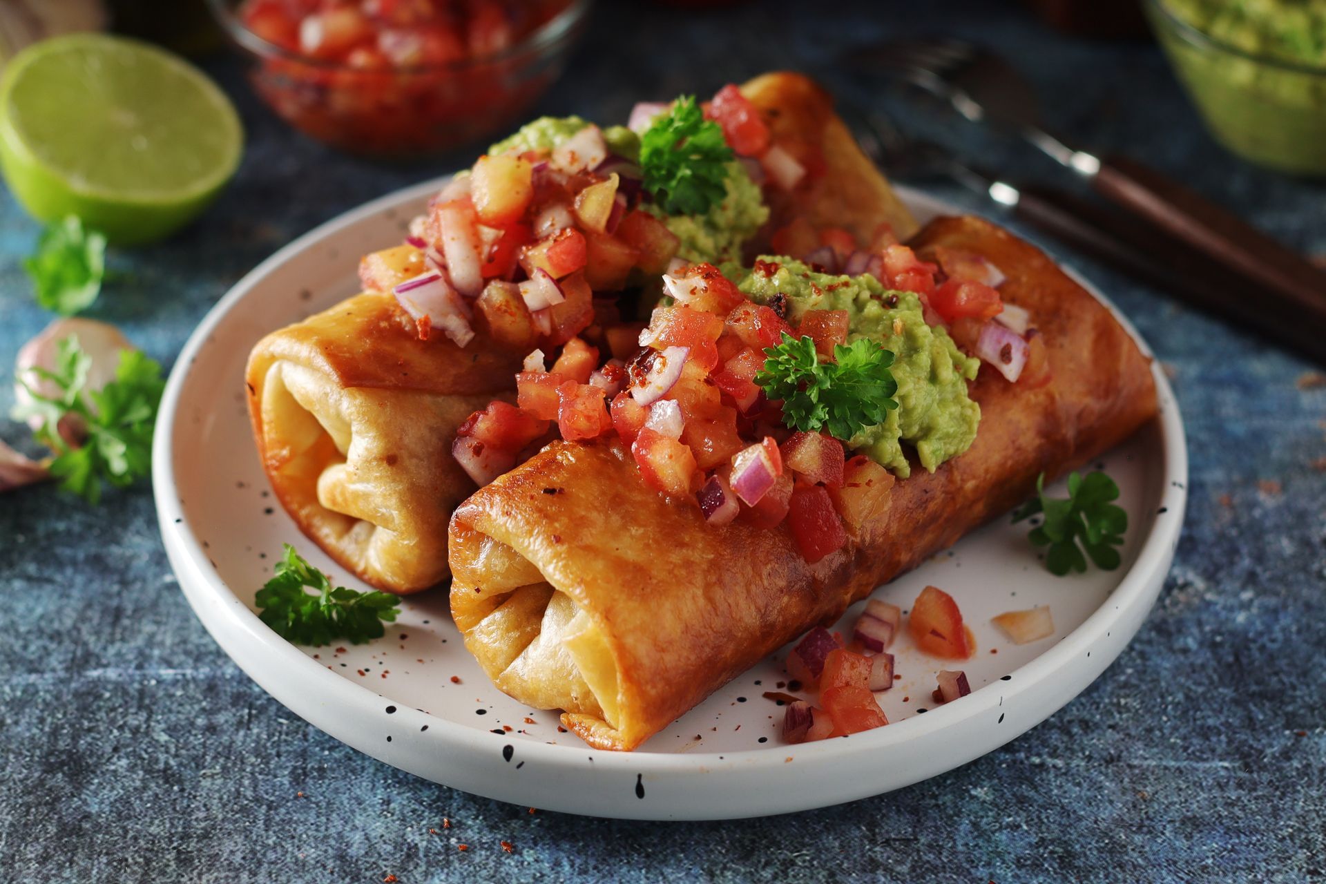Two golden-brown chimichangas topped with guacamole, fresh pico de gallo, and parsley on a white plate.