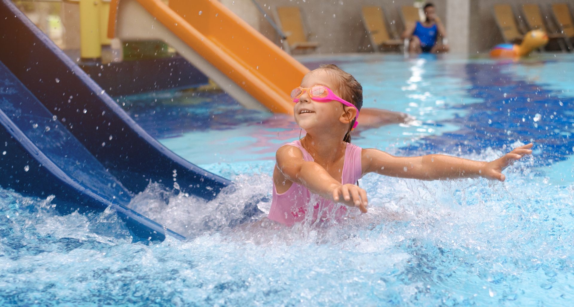 A child wearing pink goggles and a swimsuit splashes in a pool at the bottom of a blue water slide.