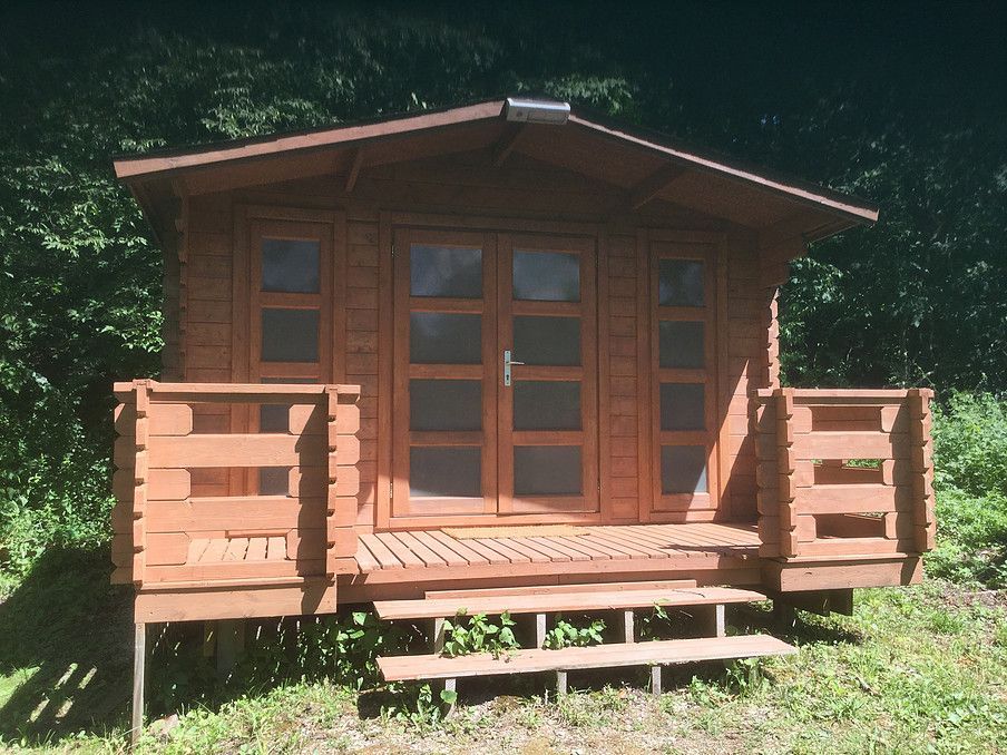 A small, brown wooden cabin with a front porch, double glass doors, and steps, surrounded by green foliage.