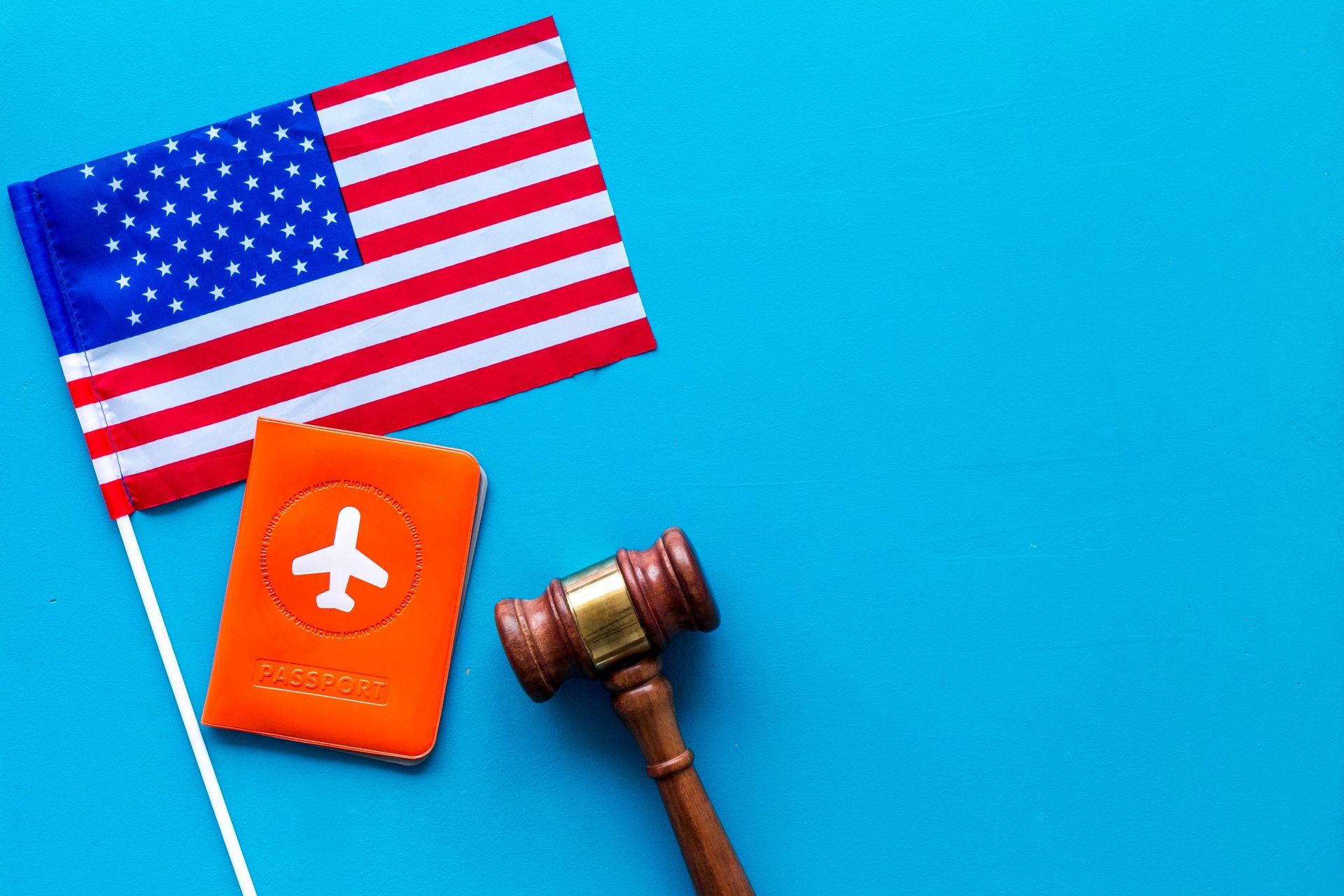 A US flag, an orange passport holder with an airplane icon, and a wooden gavel, arranged on a bright blue background.