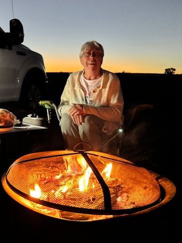 A person sits behind a glowing fire pit at dusk, with the silhouette of a vehicle nearby.