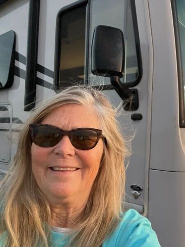 A smiling person wearing sunglasses in a light blue shirt, standing in front of the side of a large recreational vehicle.