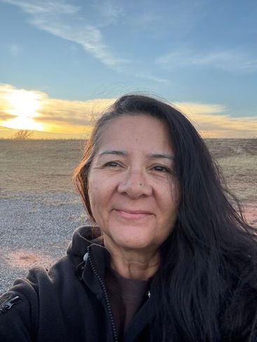 Person smiling outdoors at sunset with a grassy field and blue sky behind them