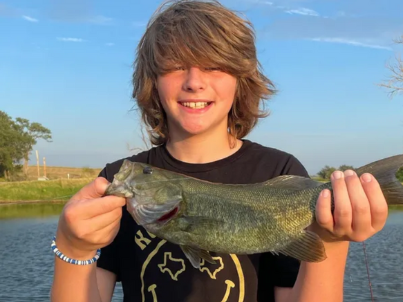 A person holding a freshly caught bass in front of a pond under a clear blue sky.