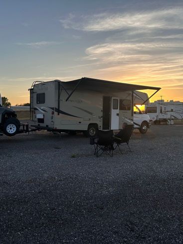 A sunset view of a recreational vehicle parked on a gravel lot with an extended awning and outdoor chairs.