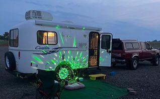 White camper with green laser lights parked beside a red truck at dusk, outdoor camping setup.