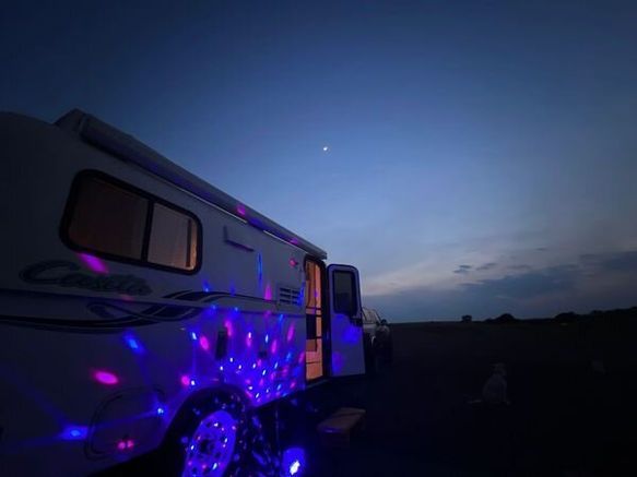 A white camper van parked at dusk, illuminated by colorful, swirling disco lights against a deep blue sky.