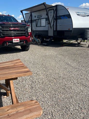 A red GMC truck parked in front of a travel trailer with an extended awning on a gravel lot near a wooden picnic table.