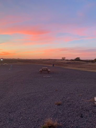 A lone picnic table sits on a gravel lot under a sunset sky filled with pink and orange clouds.