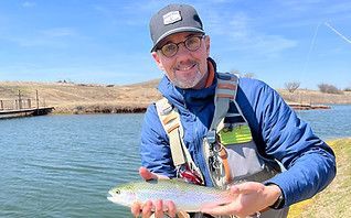 Man holding a rainbow trout on a boat, wearing a blue jacket and cap on a calm lake.
