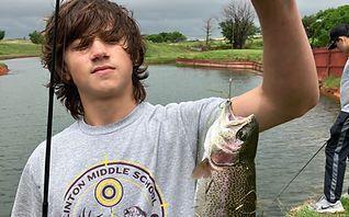 Teen holding a freshly caught fish by a pond, wearing a gray T-shirt under cloudy skies