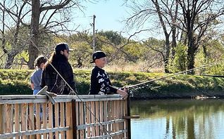 Three people fishing from a wooden dock beside a calm pond on a sunny day