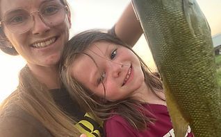 Smiling woman and child posing with a large fish outdoors at sunset