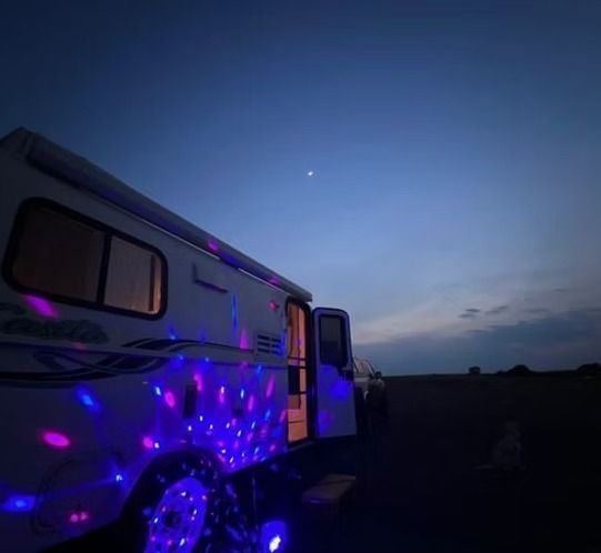 A camper van parked in a dark field at dusk, illuminated by vibrant blue and pink disco lights reflecting on its side.