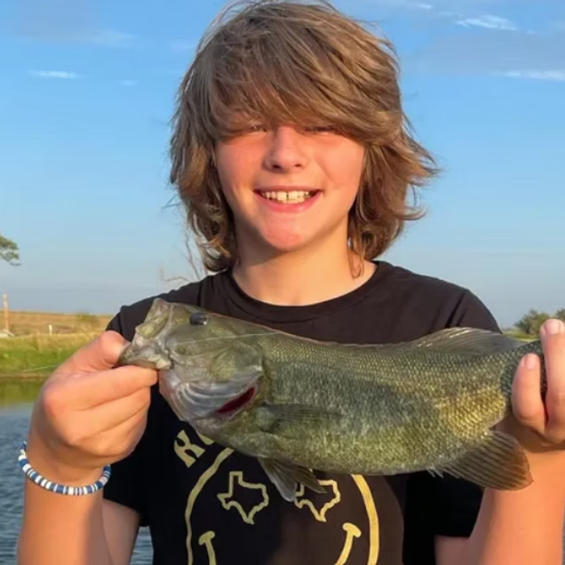 A smiling person holds a freshly caught green bass fish outdoors in front of a blue sky and water.