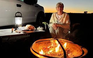 Person sitting by a campfire beside a white SUV at dusk, with a lantern and picnic table nearby