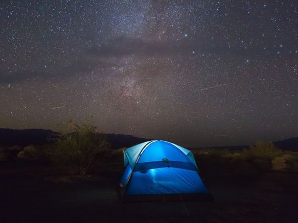 A blue tent illuminated from within, set against a dark landscape under a starry night sky with the Milky Way visible.