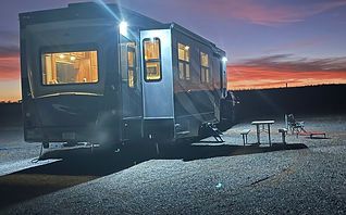 A silver trailer lit at dusk on a paved lot, with picnic tables and a sunset sky in the background
