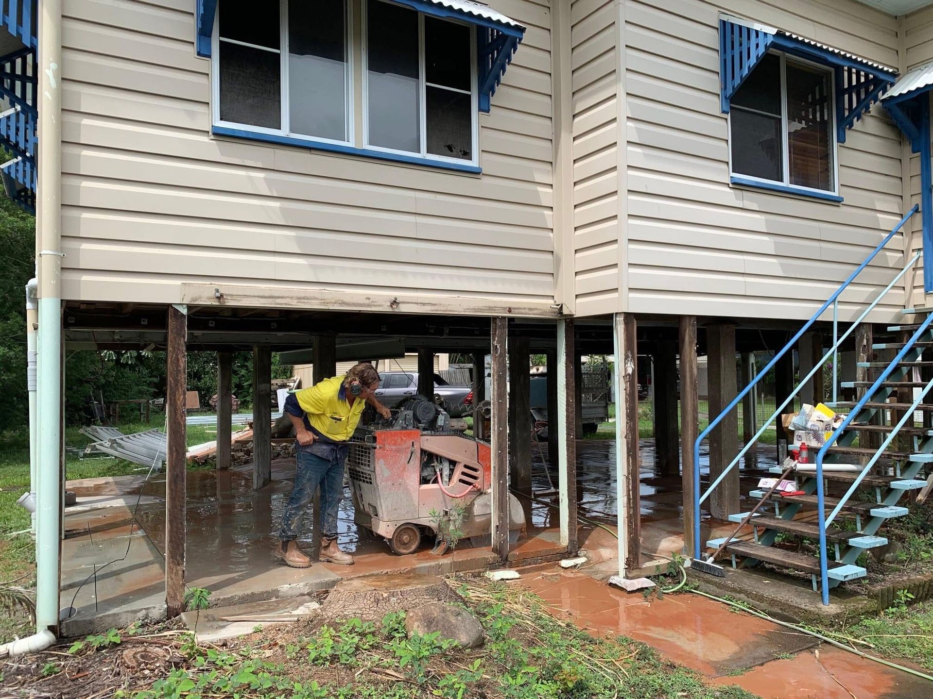 Elevated house on stilts with stairs, a person in yellow by the underside, and a cement mixer below. — Ablecut Concrete Cutting & Coring In East Mackay, QLD