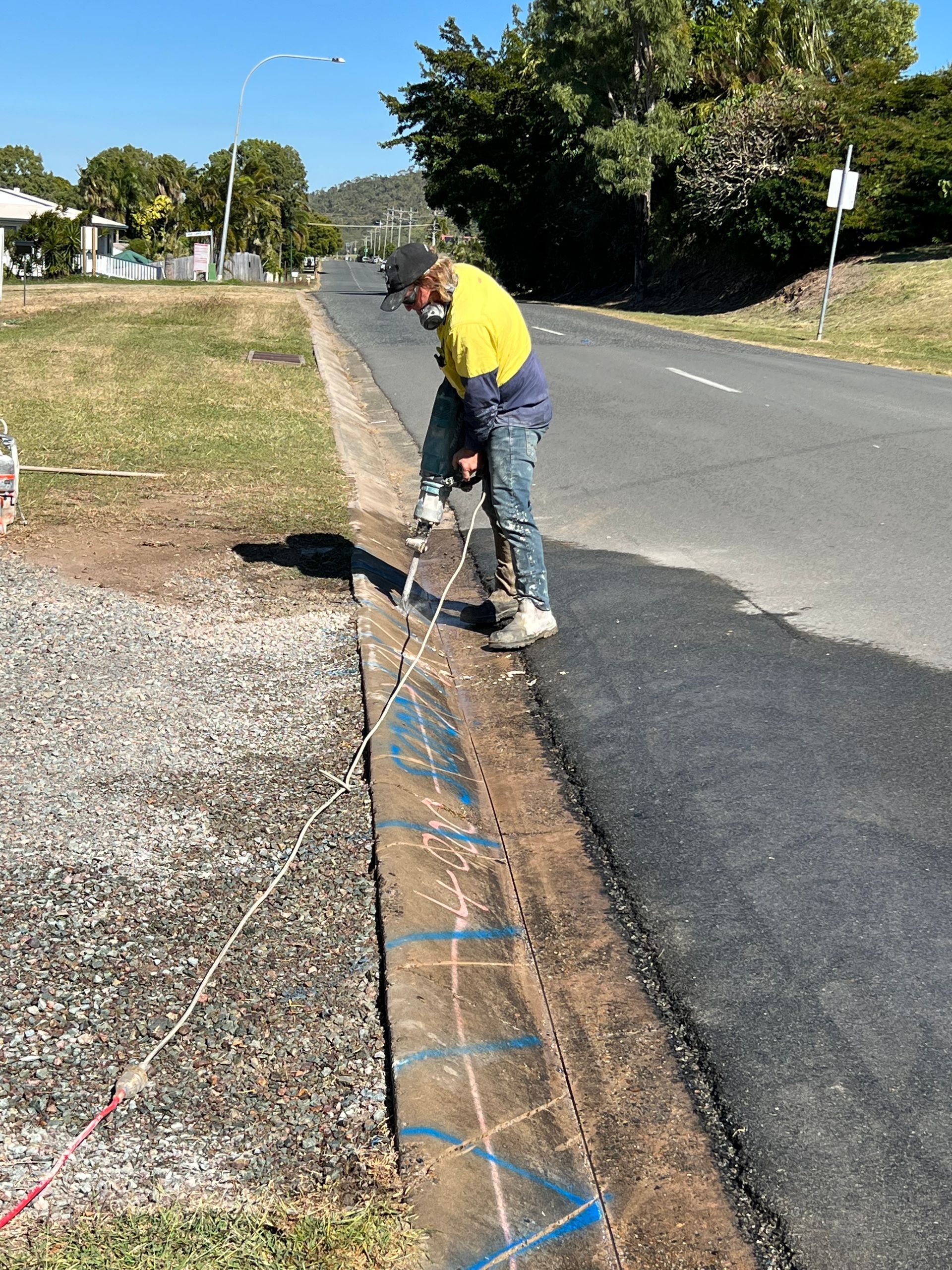 Worker in yellow shirt laying utility line along roadside trench with gravel and blue markers — Ablecut Concrete Cutting & Coring In East Mackay, QLD