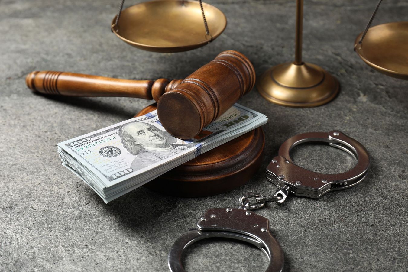 Gavel, scales of justice, stack of money, and handcuffs on a gray surface, representing justice and legal proceedings.