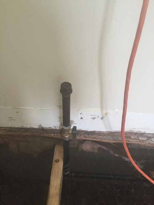 A vertical black gas pipe with a shut-off valve emerges from the floor near a wall.