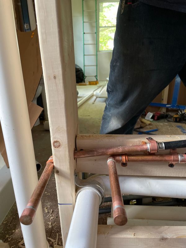 Plumbing work in progress. Copper pipes and PVC pipes are mounted in a wood frame, with a person visible in the background.