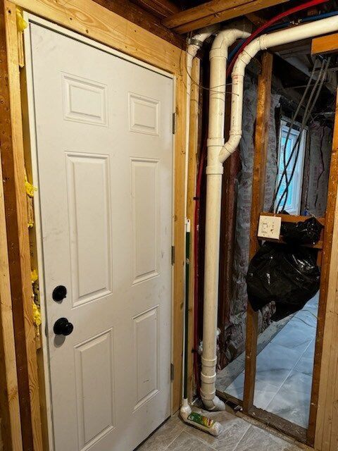 White door in a wooden frame. PVC pipes run alongside the door, with a glimpse of a dark room to the right.