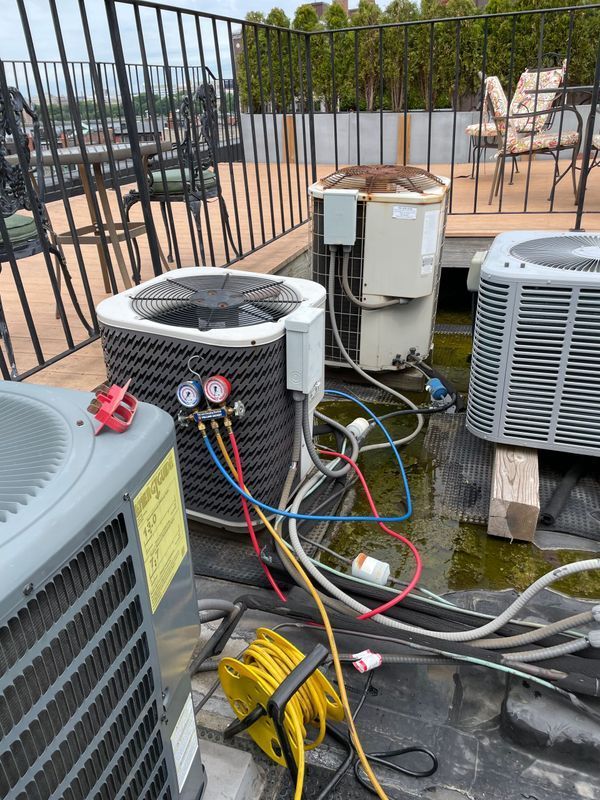HVAC units on a rooftop. Gauges, hoses connected to a unit; yellow extension cord; fencing, patio furniture in background.