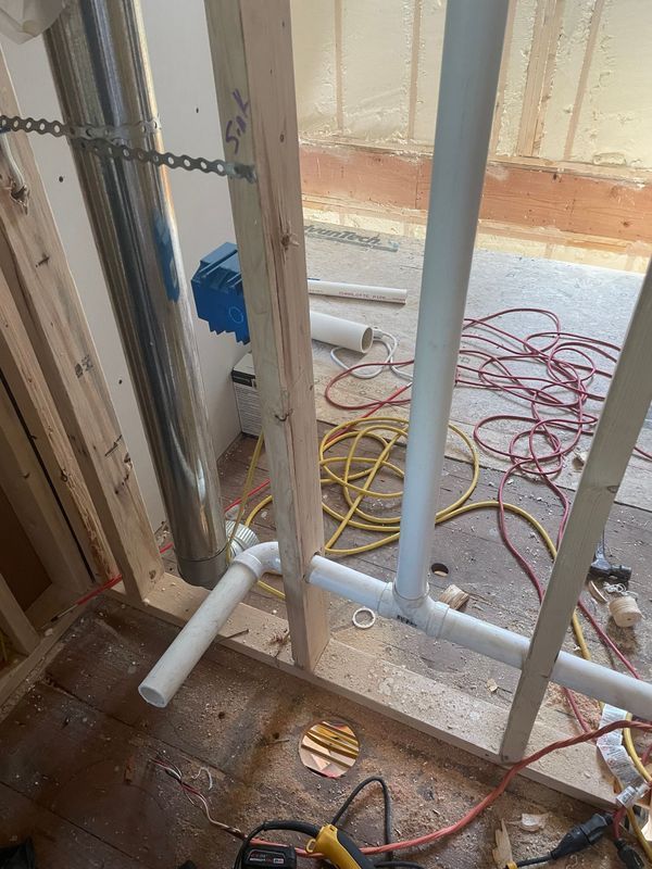 Plumbing pipes and a metal chimney in a partially constructed wall. Wiring and tools on the floor.