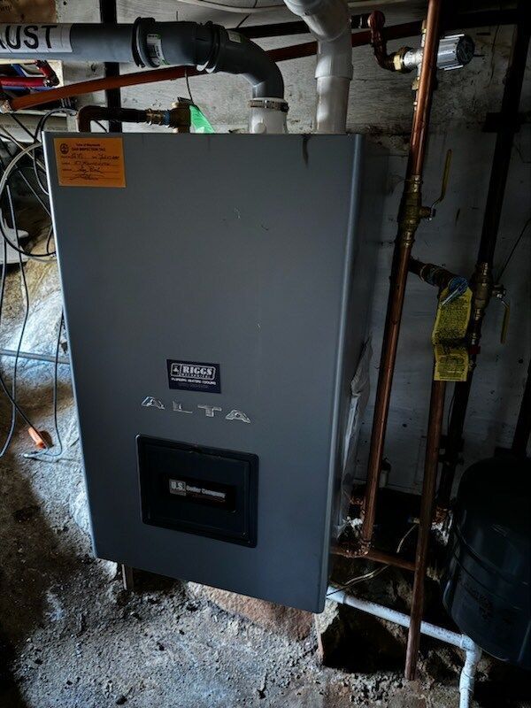 Gray wall-mounted boiler in a basement, with copper pipes and a black vent.