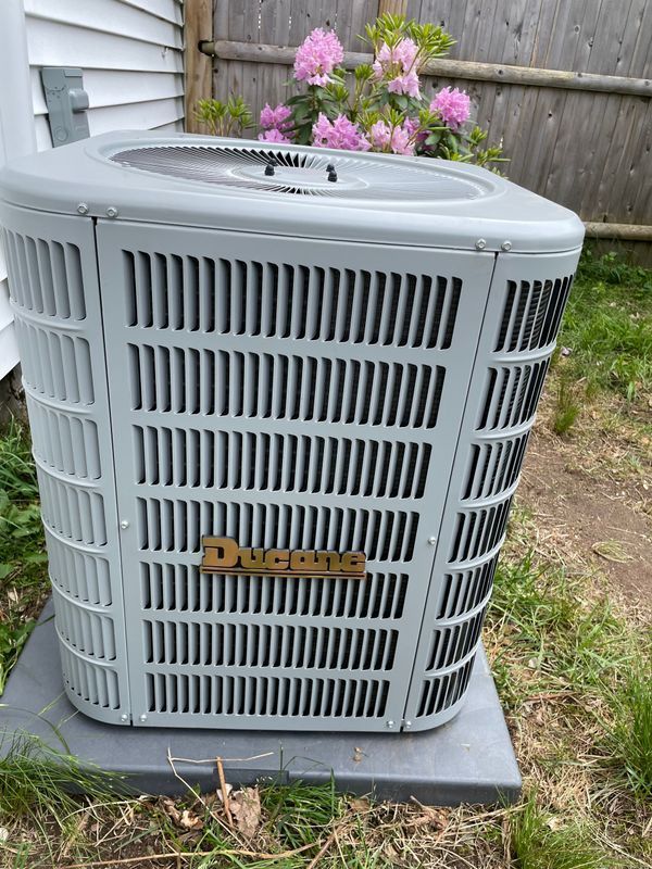 Gray air conditioning unit outside a white house, with pink flowers in the background.