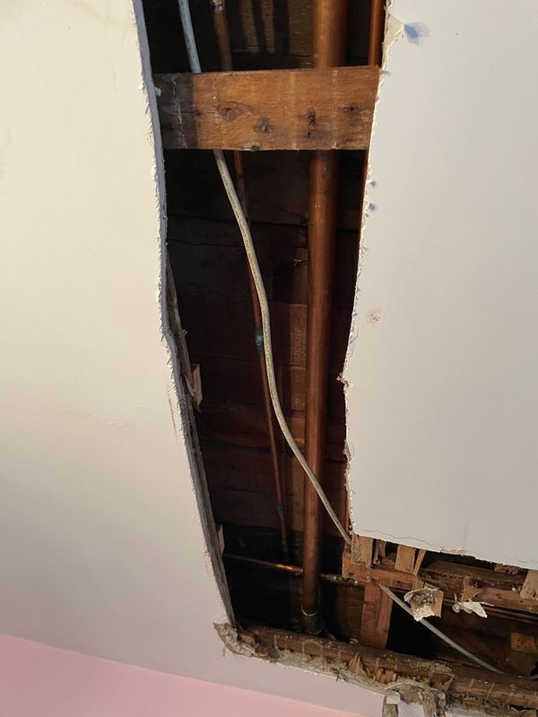 Hole in ceiling reveals wood beams, copper pipe, and electrical wiring. Drywall is cracked around the edges.