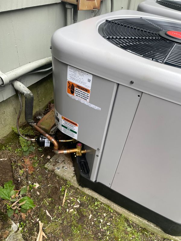 An outdoor air conditioning unit next to a light-colored building wall. Copper pipes and a black fan visible.