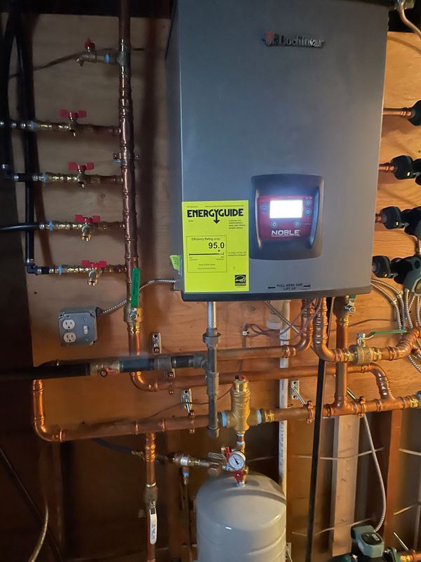 A wall-mounted, gray, Buderus boiler with copper pipes, valves, and an expansion tank in a utility room.