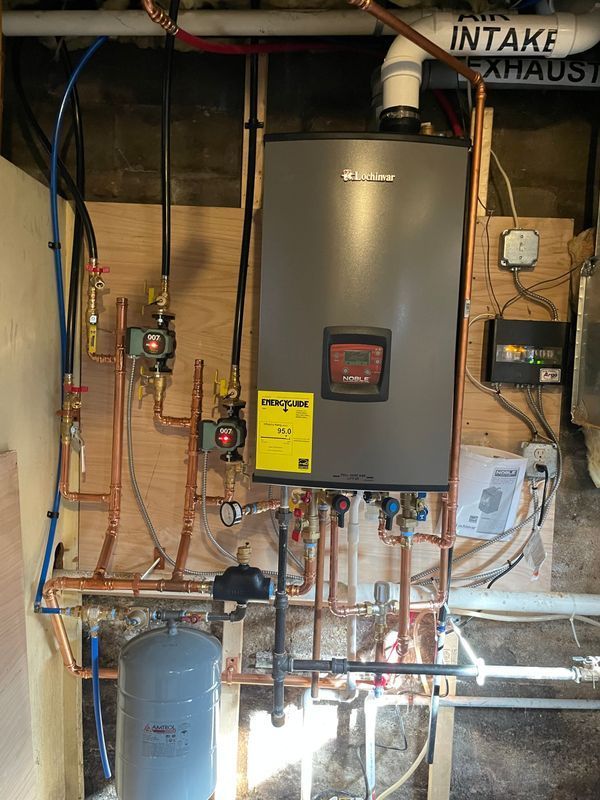 A wall-mounted gray water heater with copper pipes and pumps against a wooden surface, in a basement setting.