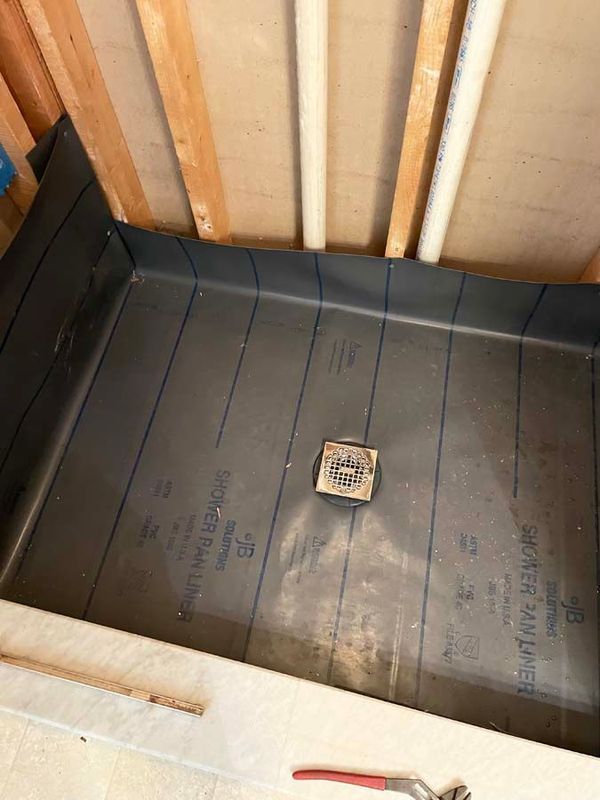 Shower pan liner installed in a framed bathroom with plumbing pipes.