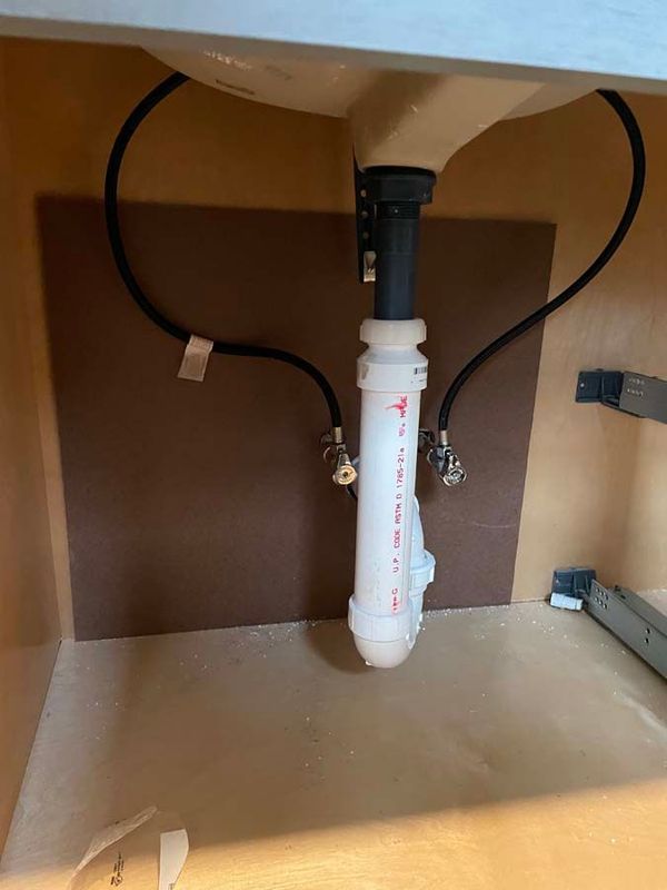 Under-sink plumbing with a white PVC pipe and black water lines against a brown backing inside a cabinet.