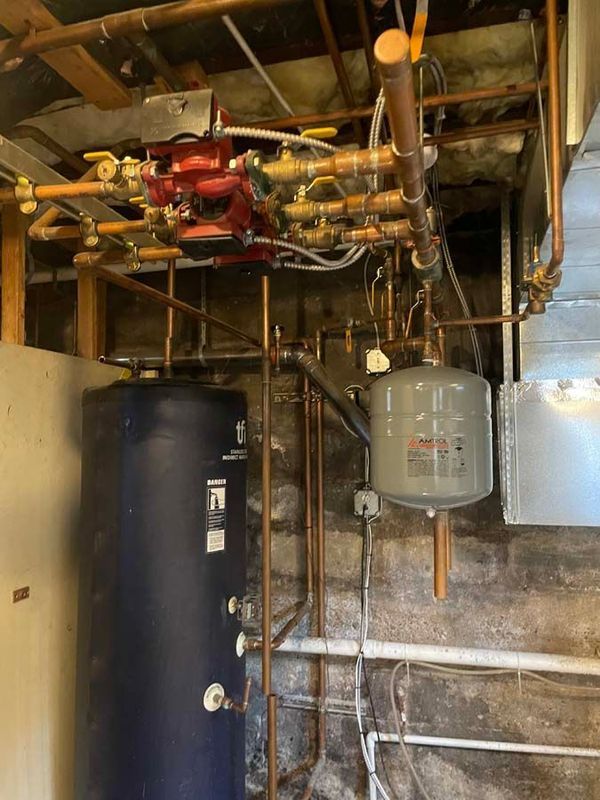 Copper pipes and red machinery connected above a blue water tank and gray expansion tank in a basement.
