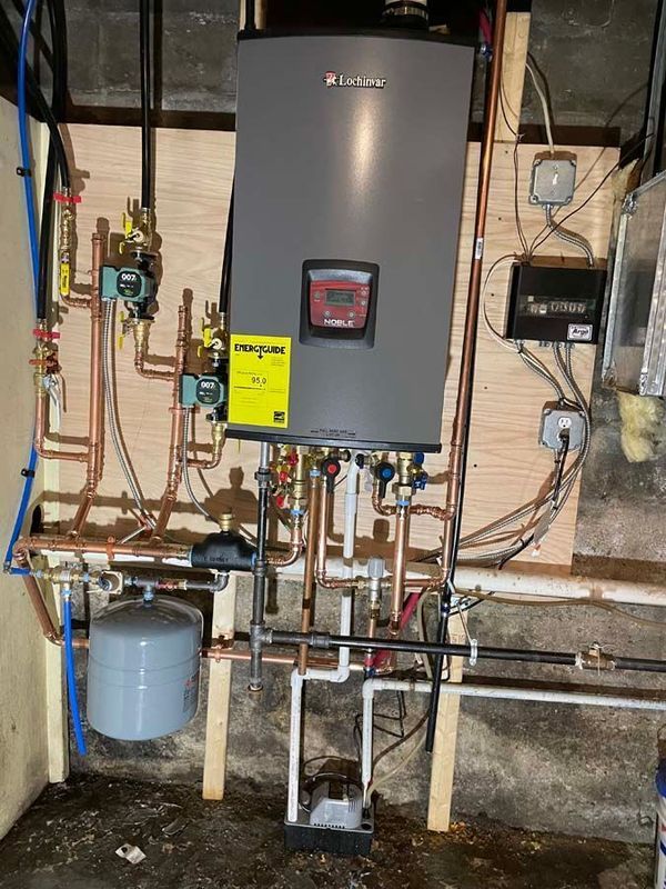 Water heater and plumbing with copper pipes on a wooden panel, against a basement wall.