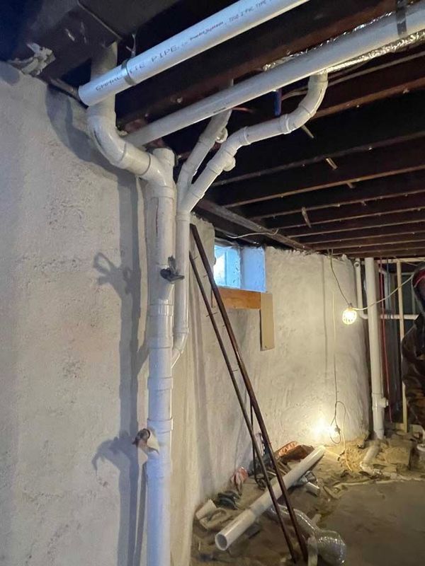 White PVC pipes against a concrete wall, basement plumbing, visible ceiling joists, and construction debris.