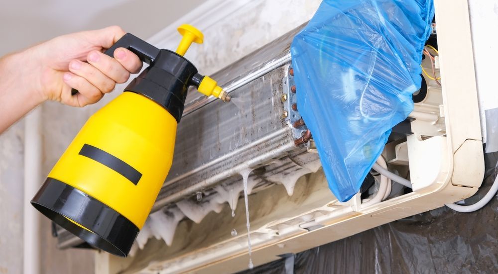 Person cleaning an air conditioner with a yellow spray bottle, foam dripping.