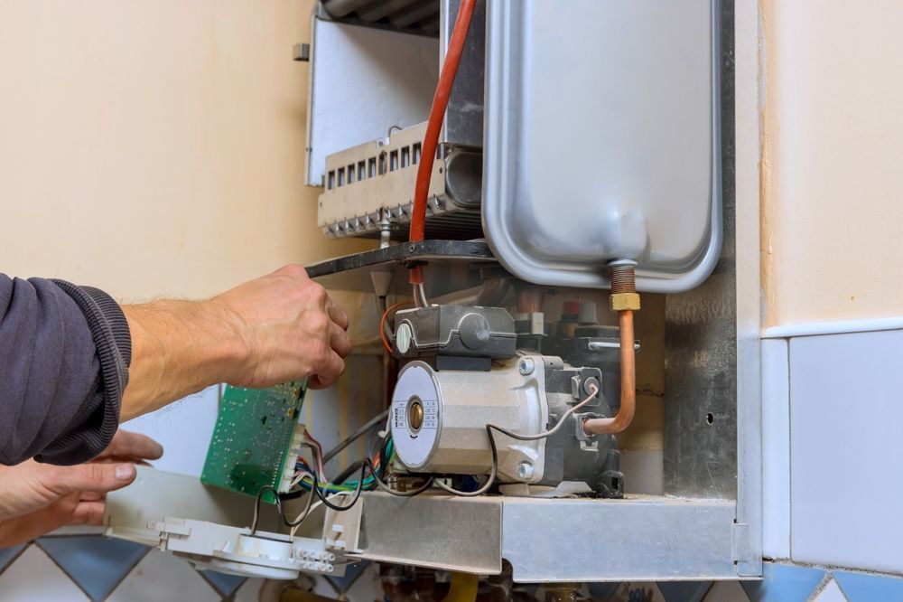Person working on a heating system. Hands reaching towards internal components, beige walls.