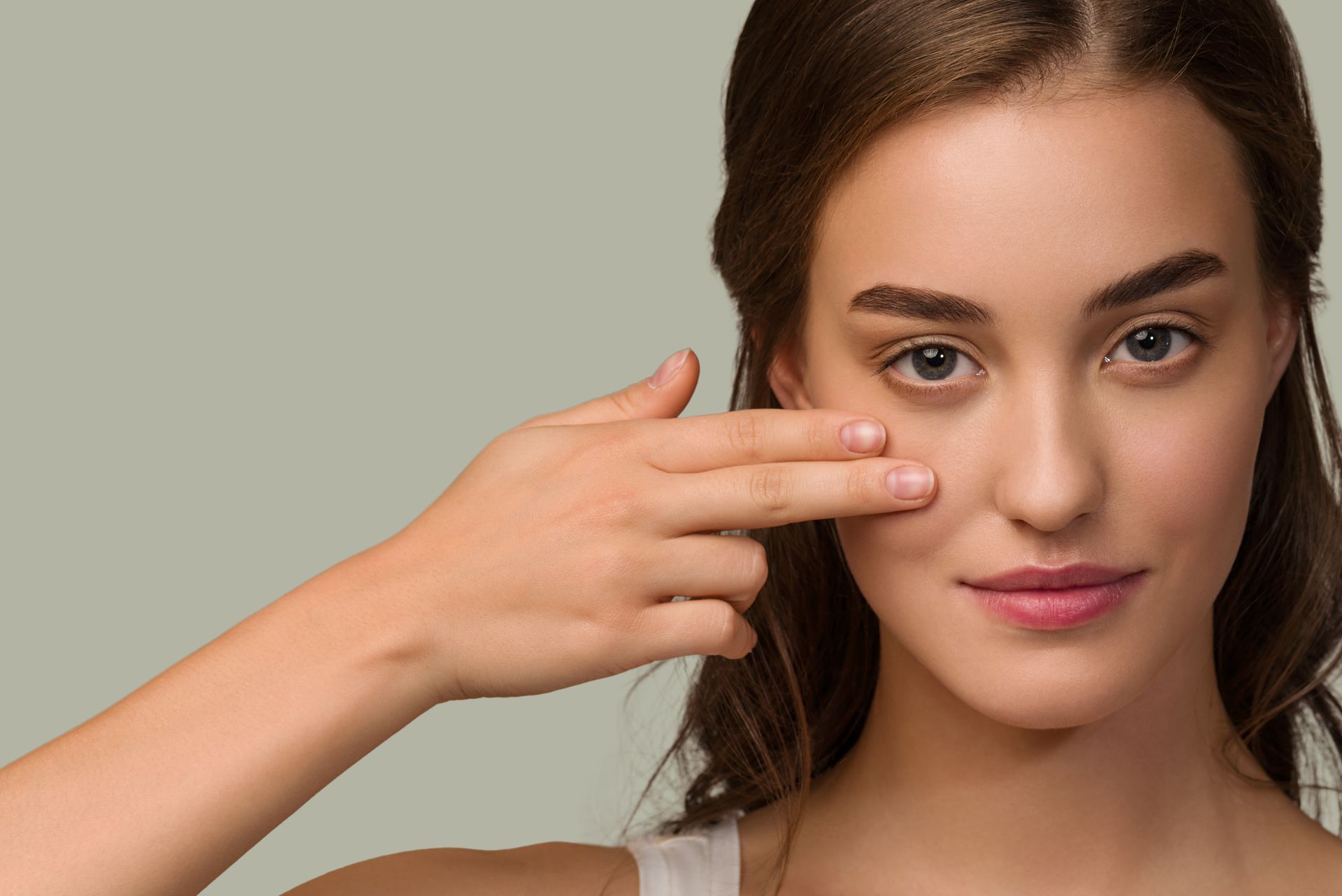 Woman with fair skin gently touches her lower eyelid, looking toward the viewer with a slight smile.
