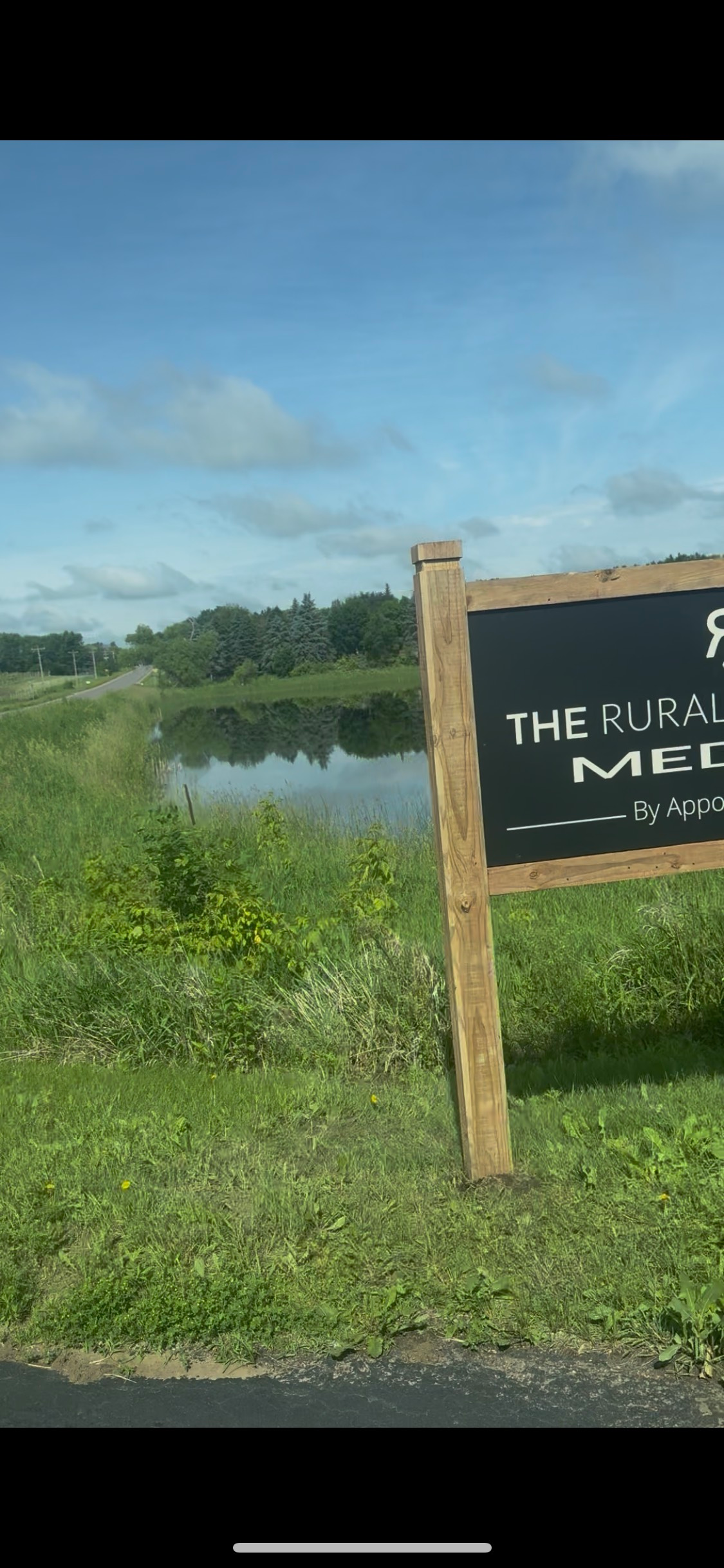 A wooden sign with text stands in front of a pond and green grass under a blue sky.