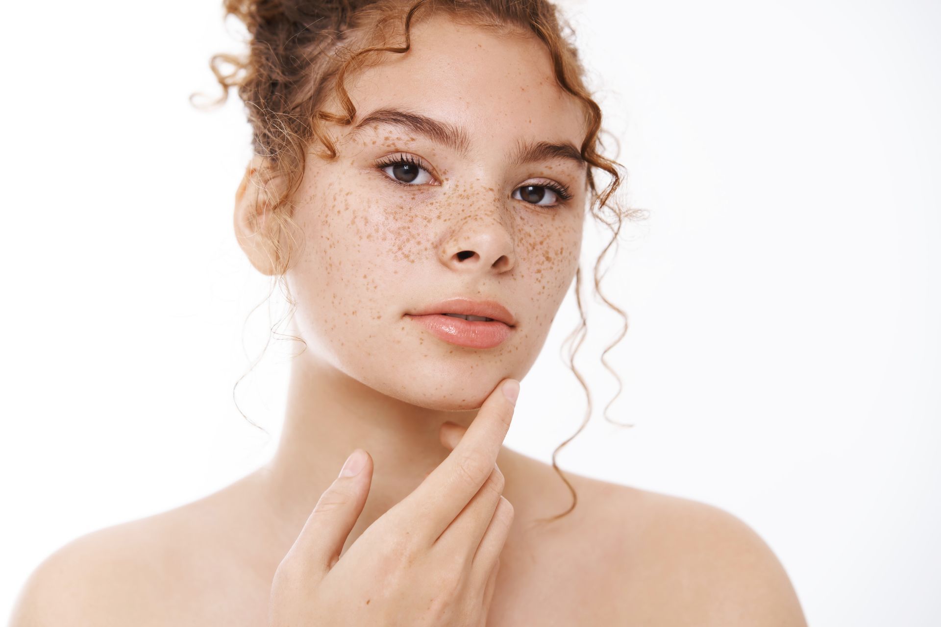Woman with freckles touching her jawline, looking at the camera against a white background.