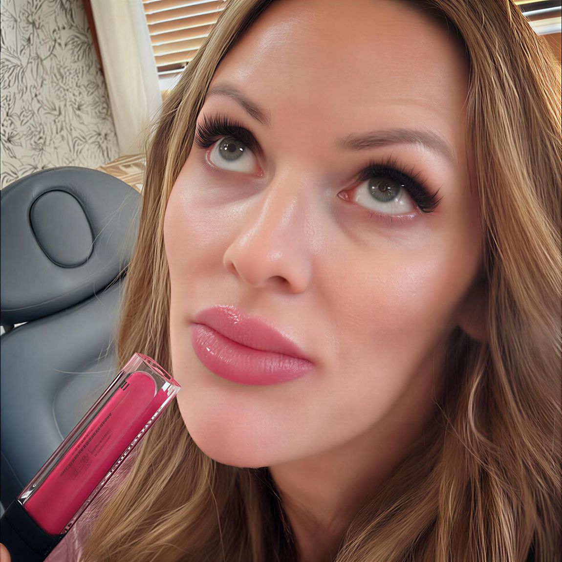 Woman looking upwards, holding a pink lipgloss tube. Her lips are glossy, and she has long eyelashes.